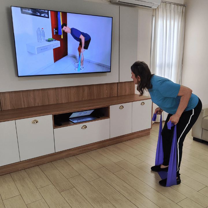 Aluna treinando com elástico na sala de casa, com o Adriano fazendo o treino junto na TV